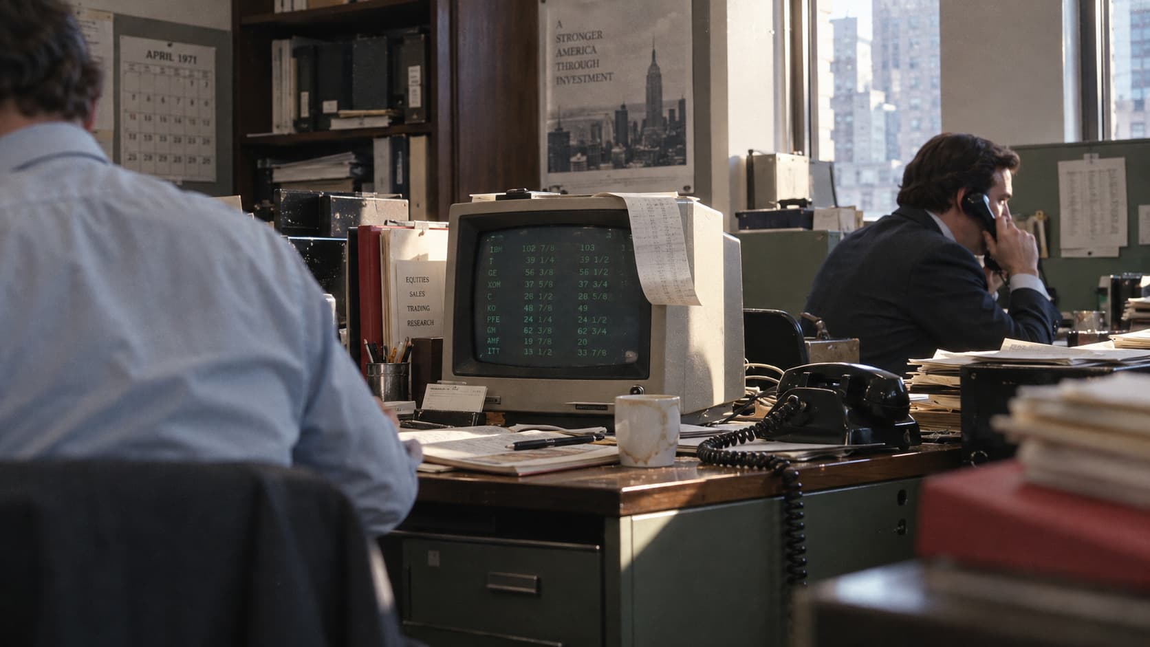A candid view of a 1971 dealing desk with a green quotation terminal, corded telephones, papers, and two dealers